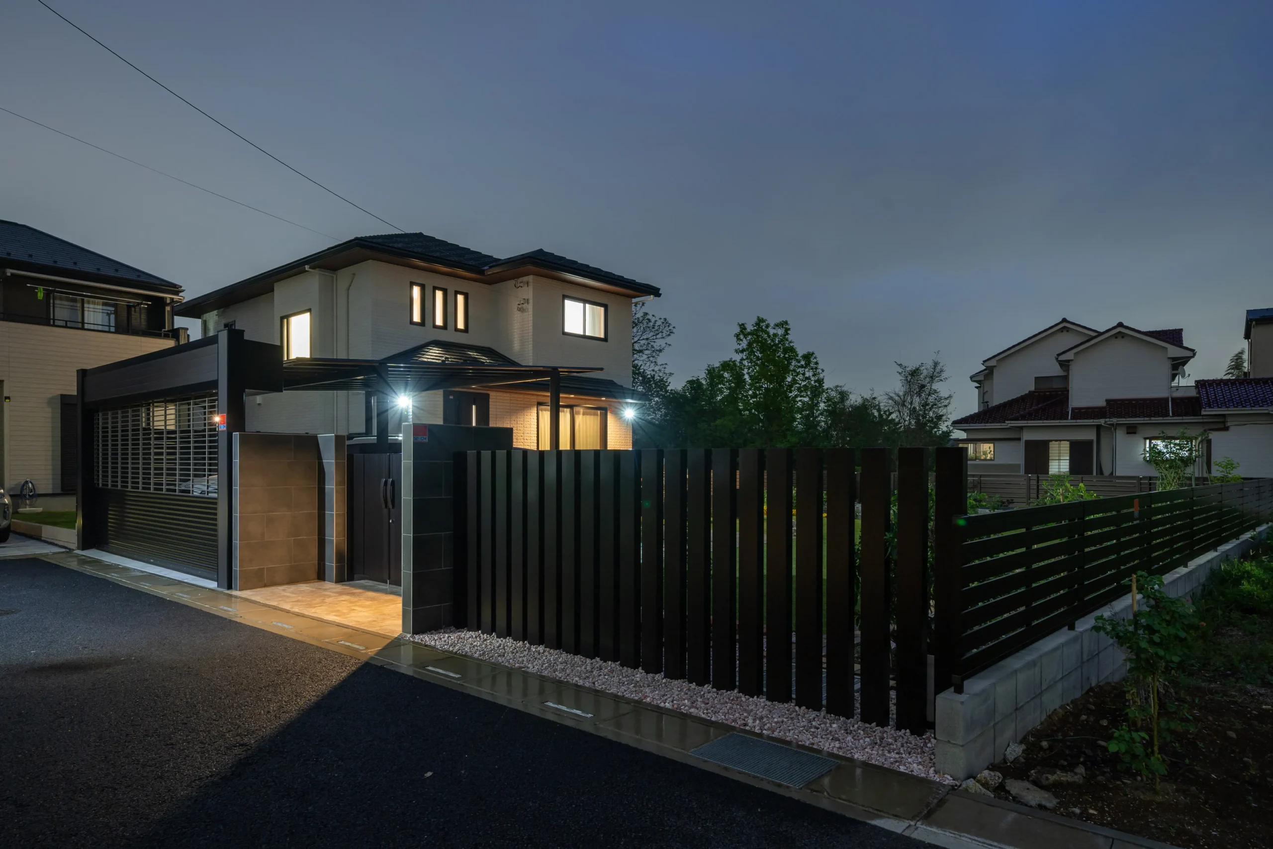 夕暮れ時のモダンな 2 階建ての家。ポーチのライトが点灯しています。家の外観は暗く、窓は大きくなっています。敷地は黒い金属製のフェンスで囲まれており、左側にはゲート付きの私道があり、フェンスの内側には砂利道があります。背景には他の家や緑が見えます。