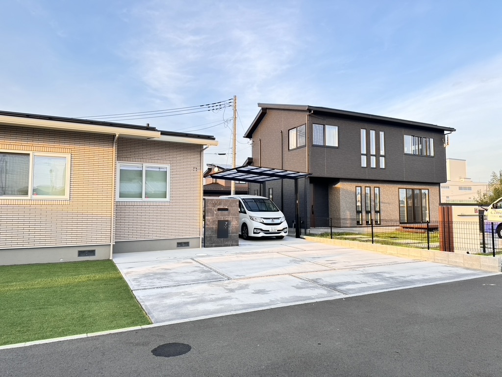 大きな窓のあるダークグレーのモダンな 2 階建ての家が、ベージュの 1 階建ての家の隣に立っています。白い車が私道に駐車されており、黒いカーポートで部分的に覆われています。前庭には小さな緑の人工芝があり、日光が差し込んでいます。.