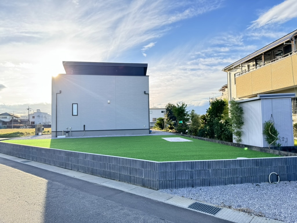 平らな屋根のモダンでシンプルな家が、手入れの行き届いた芝生のある角地に建っています。太陽は建物の背後に沈み、暖かい光を投げかけています。空は部分的に曇り、敷地の横には舗装道路が走っており、その周囲には歩道と砂利のエリアがあります。.