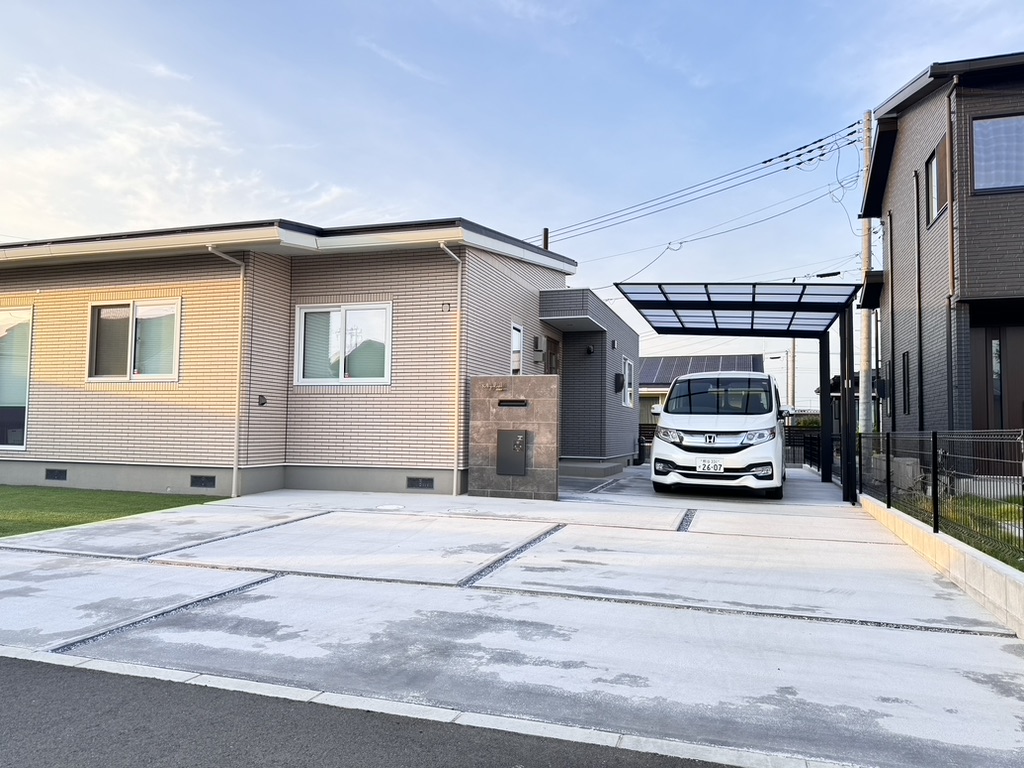 石で縁取られた正面と右側に車道がある、モダンな平屋建ての家。黒いカーポートの下に白いバンが駐車されています。車道はコンクリートの板で縁取られています。空は晴れていて、少し雲がかかっており、車道の左側には芝生があります。.