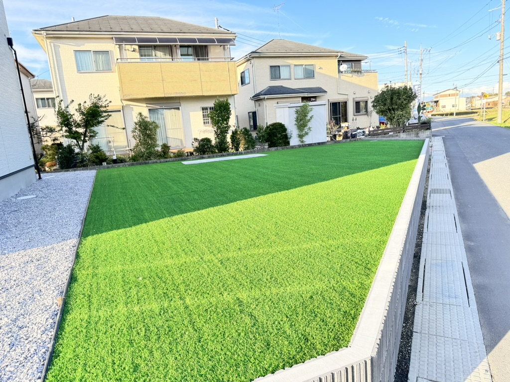 大きな窓とベージュの外装を備えた 2 軒のモダンな 2 階建ての家が特徴の住宅街。手前には青々とした人工芝があり、右側には狭いコンクリートの小道が続いています。空は澄み切った青空で、晴れた日を予感させます。.