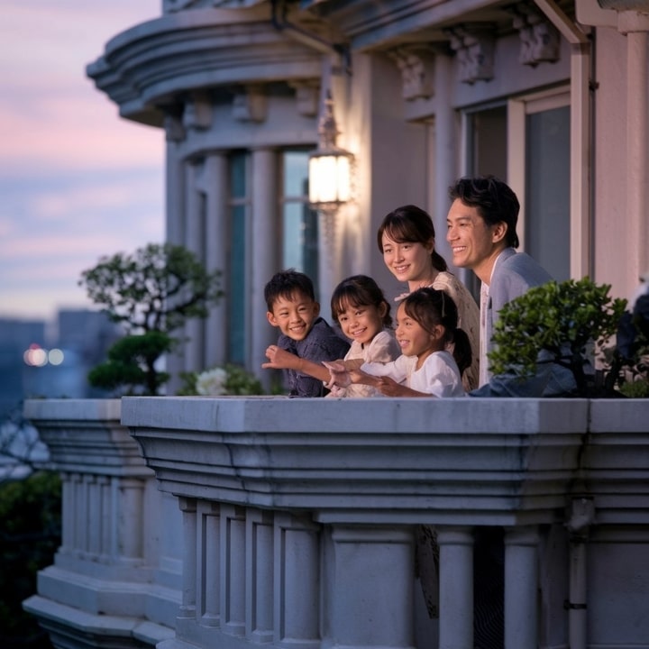 上尾市の夕暮れ時、5人家族がバルコニーに立っています。両親と3人の子供たちが楽しそうに外を眺めています。2人の娘は白いドレスを着ており、息子は青いシャツを着ています。外構工事の専門知識を活かして設計されたバルコニーには、鉢植えやクラシックな建築ディテールが置かれ、温かい雰囲気を醸し出しています。