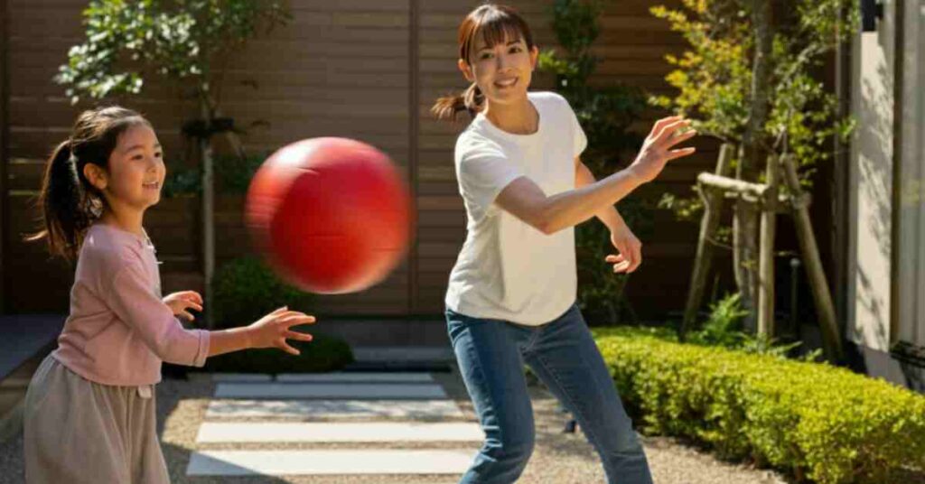 女性と少女が庭で大きな赤いボールで遊んでいます。白い T シャツとジーンズを着た女性はボールをキャッチしようとしており、ライトピンクの長袖シャツとグレーのパンツを着た少女は笑顔でボールに手を伸ばしています。緑の低木と木製の柵が、この楽しいシーンを縁取っています。