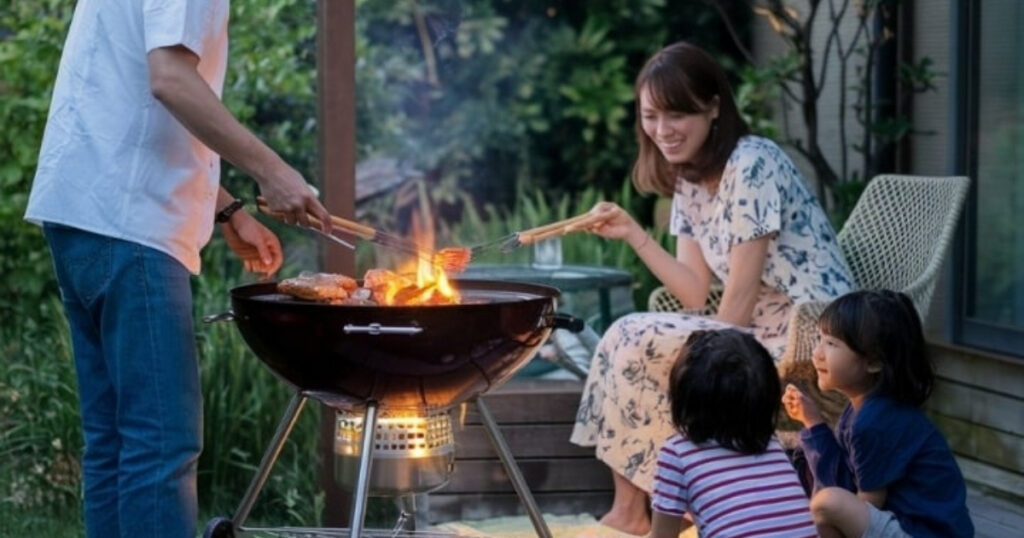 バーベキューグリルで食べ物を焼いている人のそばに、花柄のドレスを着た女性と、ストライプのシャツを着た子供 2 人と青い服を着た子供 2 人が座っています。彼らは木や茂みを背景にした芝生のエリアに集まっており、屋外での食事の体験をより一層盛り上げています。