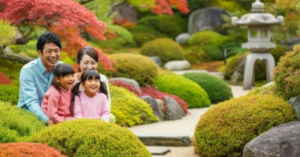 埼玉県の日本庭園にある、大人2人と子供2人の4人家族が、青々と手入れされた低木に囲まれた石畳の道に座っている。大人たちは子供たちの後ろに立ち、皆笑顔で、紅葉の穏やかで色鮮やかな景色を楽しんでいるように見える。