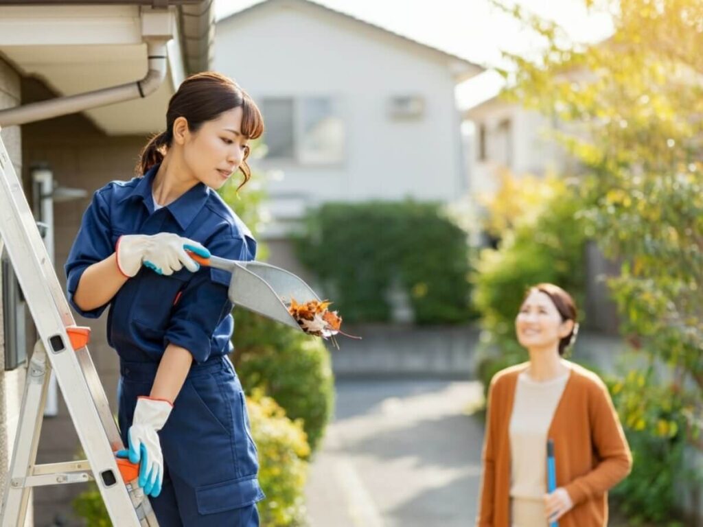 青いオーバーオールを着た人が梯子の上に立ち、葉っぱを詰めたスコップで側溝を掃除している。カジュアルな服を着た別の人が梯子の上にいる人を見上げて微笑んでいる。彼らは家族の家の近くの屋外にいて、背景には木々と家が見える晴れた日である。