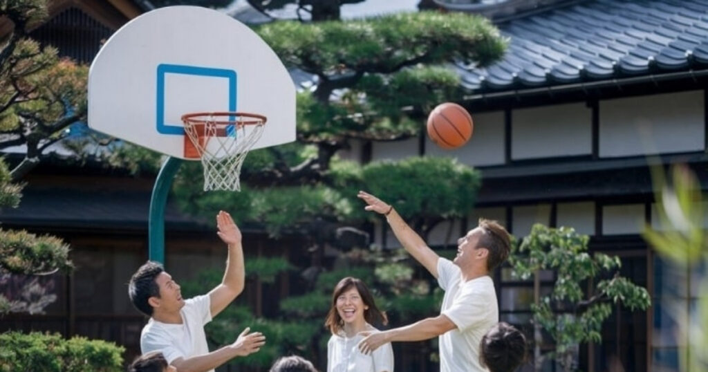 3 人の若者のグループが、伝統的な日本家屋の近くの屋外でバスケットボールをしています。1 人がジャンプしてシュートをブロックし、もう 1 人がシュートを打とうとしています。背景の女性は、豊かな緑と木々の下書きを背景に昔の記憶が蘇るのを見ているかのように、微笑みながらその様子を見ています。